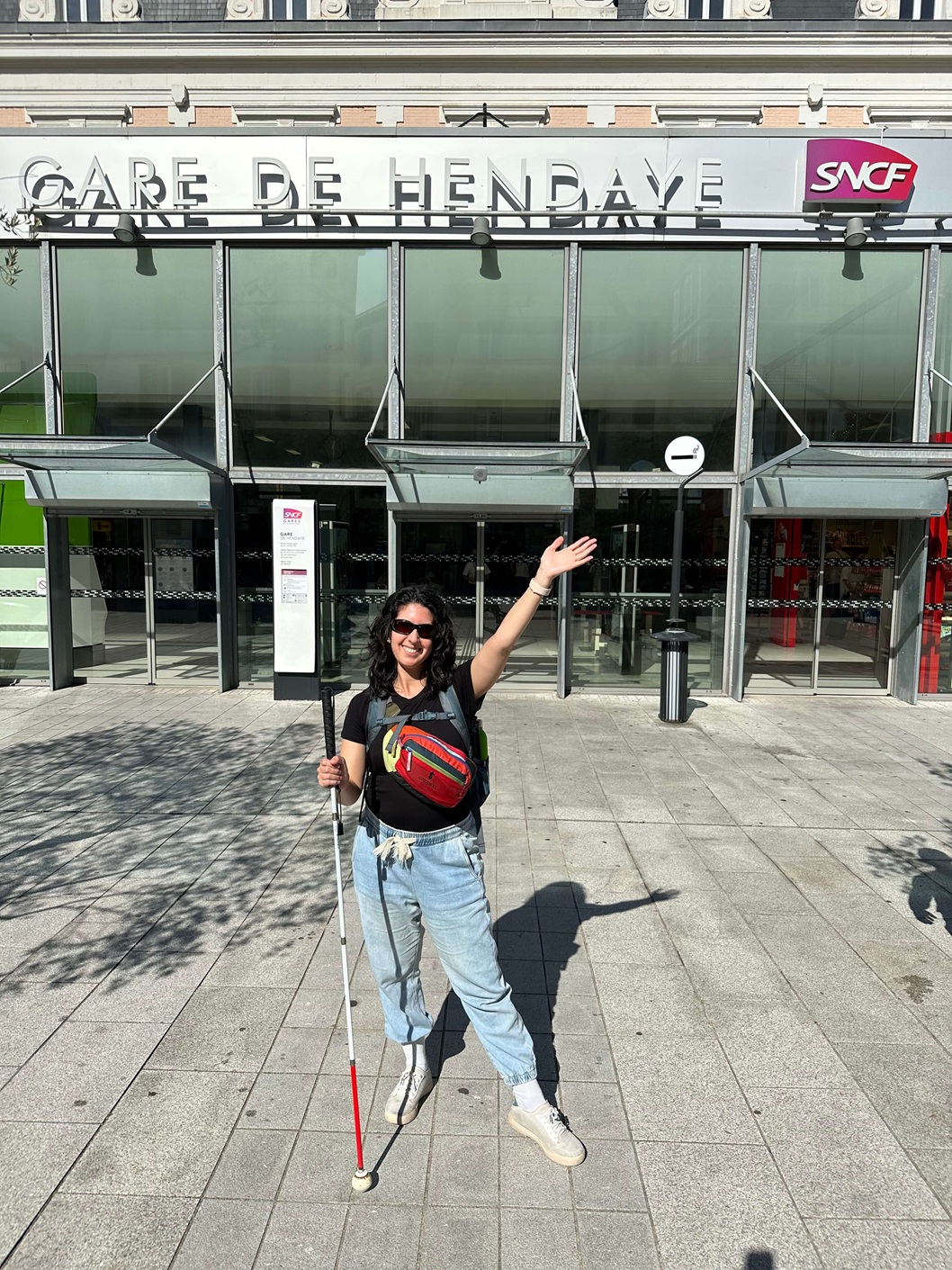 기차역 밖에서 흰색과 빨간색 지팡이를 든 여성이 'Gare de Hendaye'라는 간판 앞에서 포즈를 취하고 있습니다