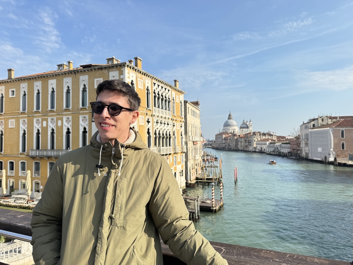 Cristian stands on a bridge with a canal and historic buildings in the background