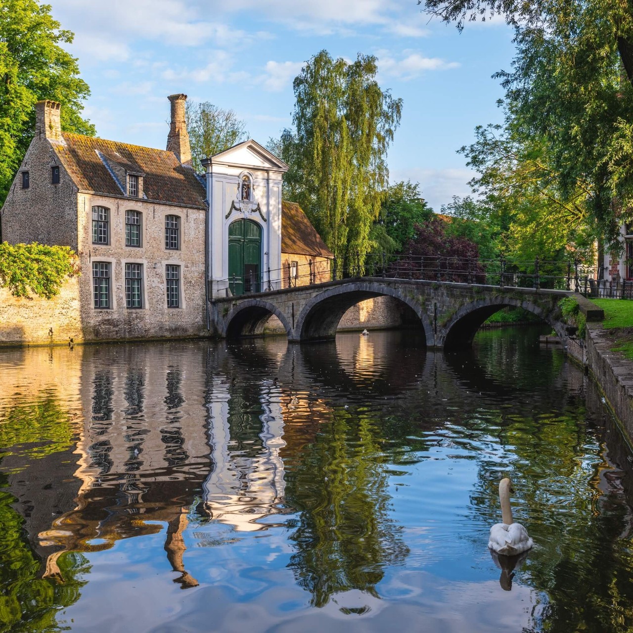 An ornate building sits on the side of a gentle river, with a bridge crossing to green grass on the opposite side