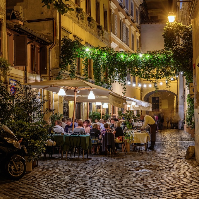 Mesas al aire libre de un restaurante, llenas de comensales, en una calle tranquila de Roma