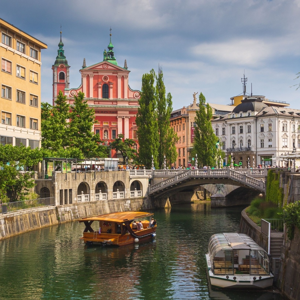 Dos barcos navegan por el río en Liubliana, con edificios históricos y un puente al fondo