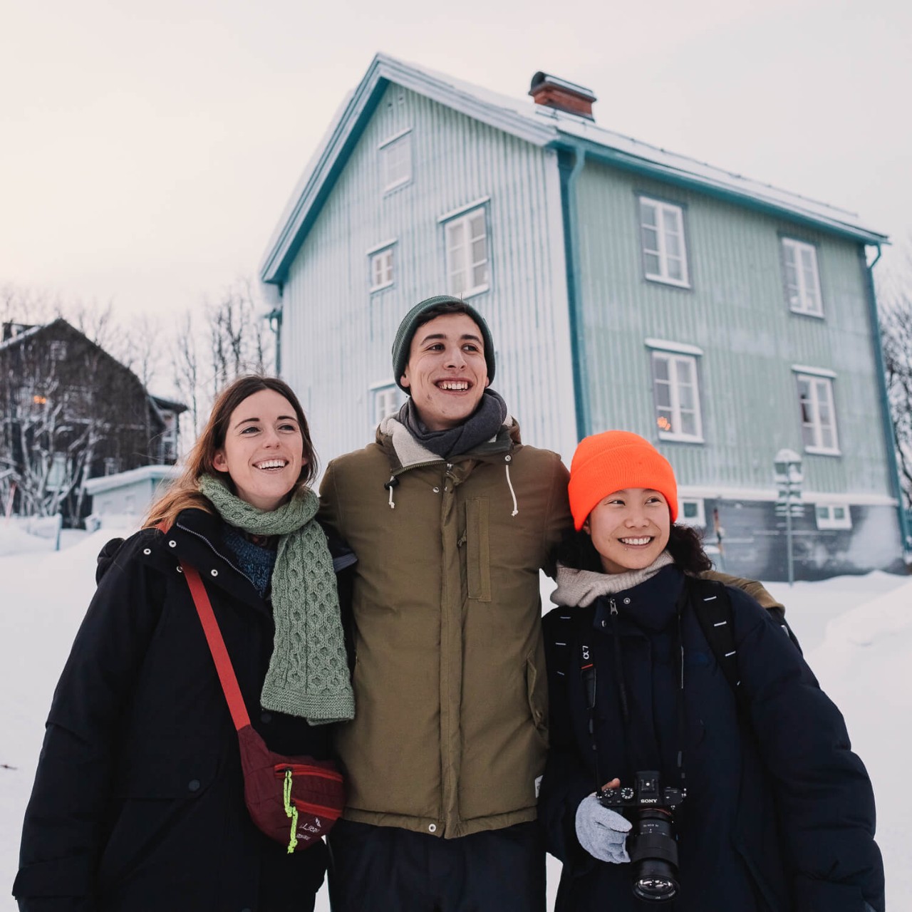 Três pessoas estão em frente a uma casa azul em uma rua coberta de neve em Kiruna, Suécia