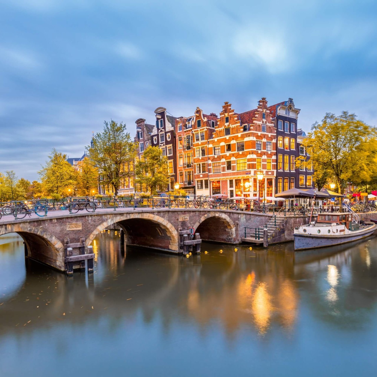 Um canal azul em Amsterdã, que passa embaixo de uma ponte, com casas altas e antigas ao fundo.